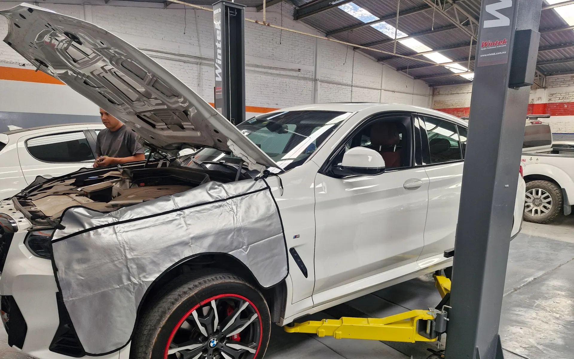 Mecánico de Motorfix trabajando en un BMW X4 blanco con el cofre abierto y fundas protectoras